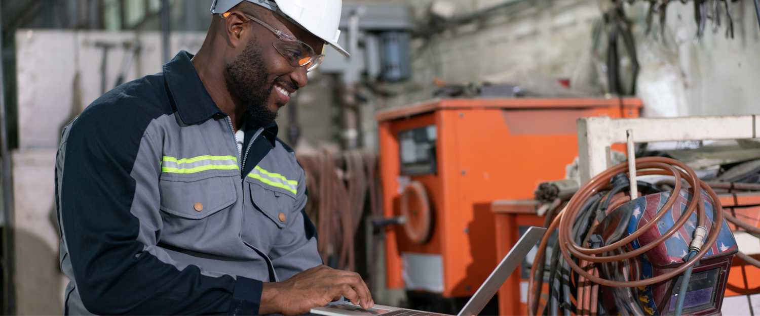 African American male engineer using computer laptop control robot arm welding machine in an industrial factory.